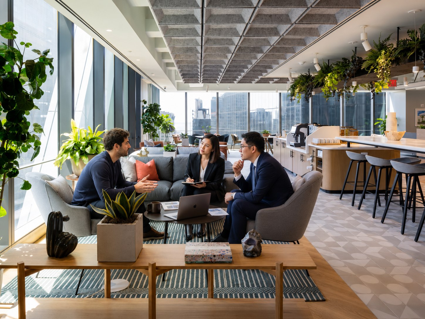 Three people in an open office lounge, one talking with his hands, one taking notes and one listening, a laptop and papers on the low table between them